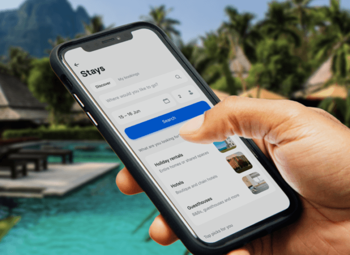 Someone is holding a phone while on holiday. The Revolut Stays app is open on the screen while a swimming pool and palm trees can be seen in the background.