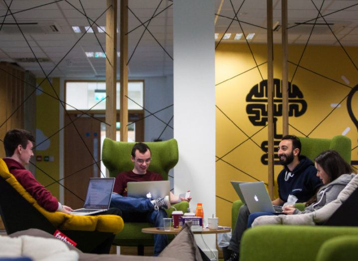 Three men and a woman sit in a round of lime-green high-backed chairs, working on laptops in a vibrant office space.