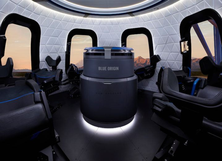 The inside of Bezos' Blue Origin spacecraft is shown. There is a central element with Blue Origin printed on it. There are six chairs around the capsule with a window behind each chair.