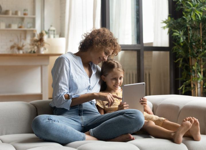 A mother is sitting with her daughter on the couch. They are smiling and playing on a tablet together.