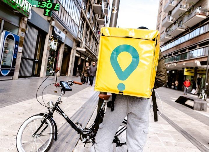 A man with a bike wearing a Glovo backpack is standing on an Italian city street.