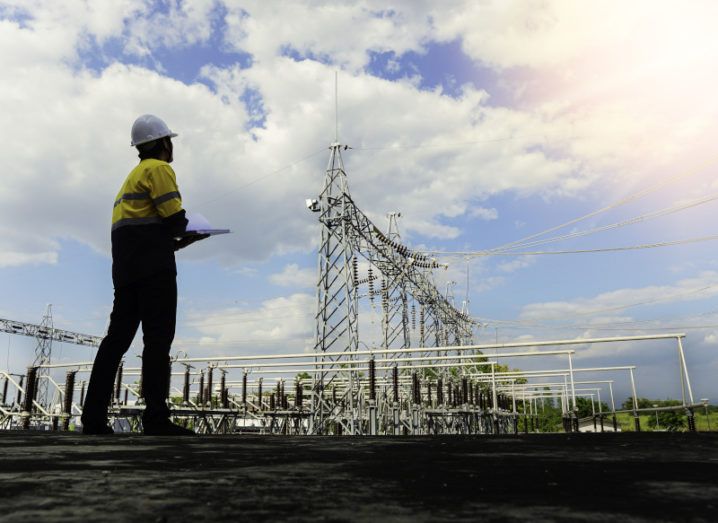 An engineer in a hard hat and a high-vis jacket looking up at a large power station against a blue sky.