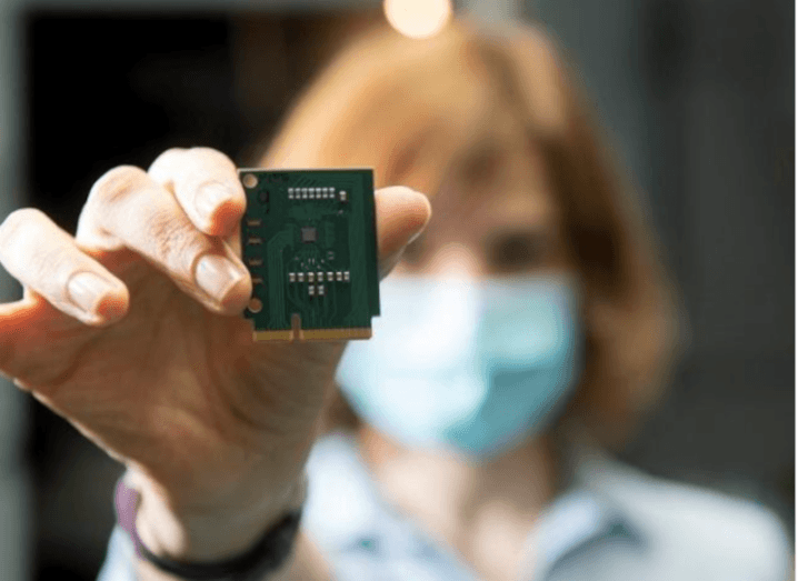 Close-up of a computer chip being held up by a woman, who is out of focus in the backgroun, wearing a face mask.