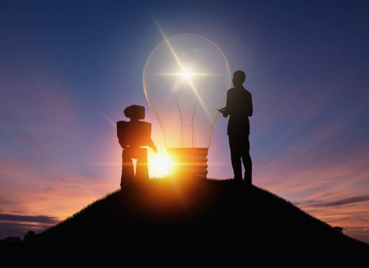 A robot and a person are standing beside each other in front of a giant lightbulb. The sun is setting in the background but the lightbulb is illuminating the surrounding area.