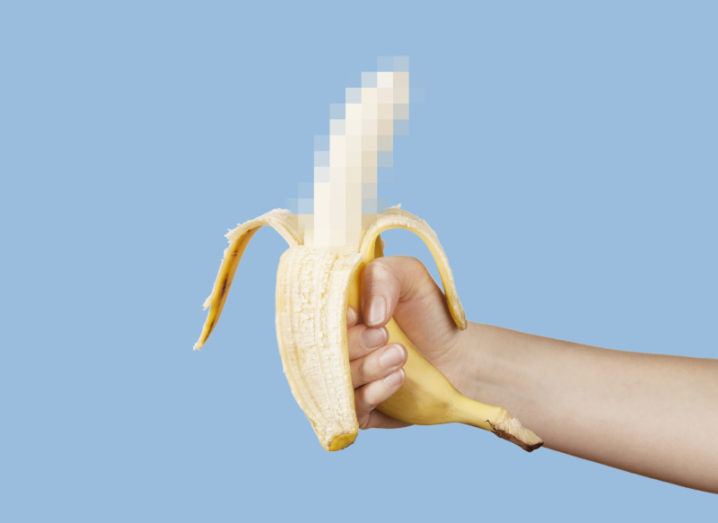 A hand holds up a banana in front of a blue background. The banana peel is stripped about halfway down the fruit, which is censored by pixelation.