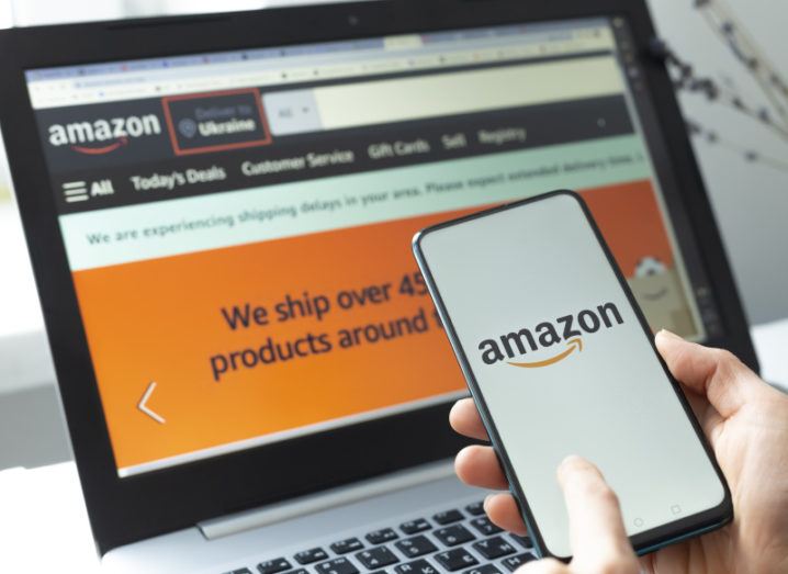 A stock image of a person holding a phone displaying the Amazon logo in front of a laptop which displays the Amazon home page.