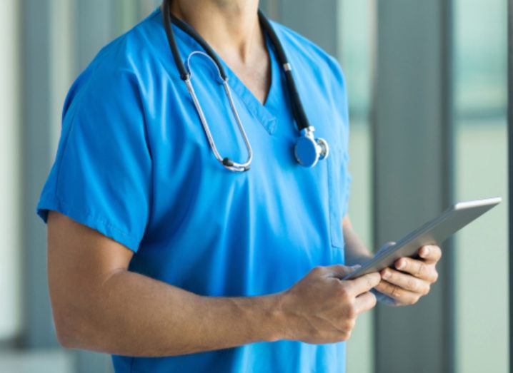 A nurse is standing in a corridor, wearing a stethoscope around their neck and holding a tablet computer.
