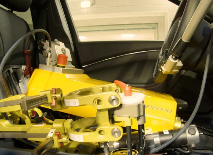 One of Ford’s yellow robot drivers can be seen sitting in the seat of the car. One arm is on the gearshift. The robot is short and squat and resembles a box on the seat, far below the height of the headrest.
