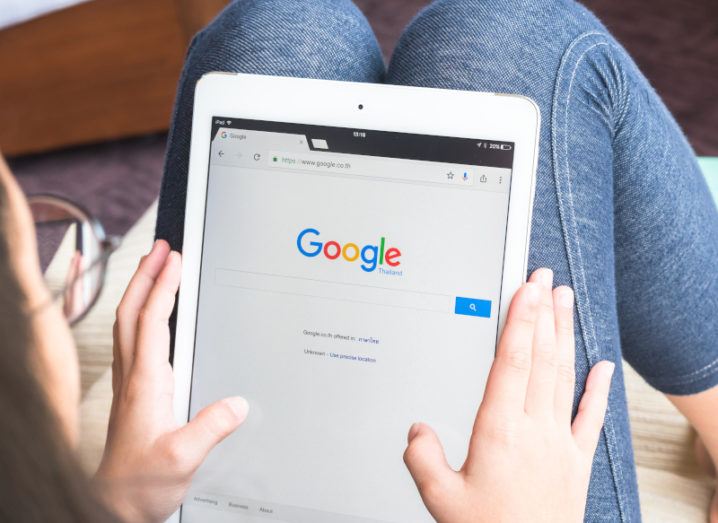 A child is holding a tablet and the Google search bar is on the screen. She is sitting down and holding the tablet against her legs while facing away from the camera.