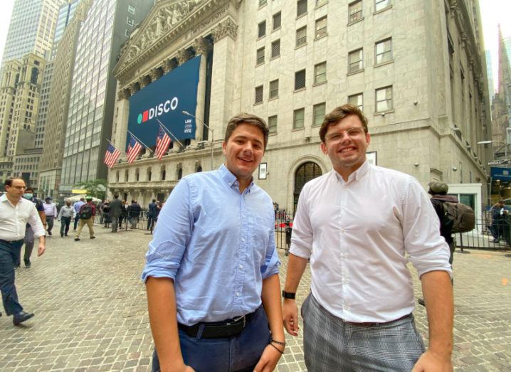 Two young men dressed in smart casual attire stand outside the New York Stock Exchange, a large neoclassical building with a facade of colonnades atop two-storey podiums.