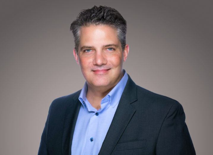 Mike Anderson, a man in a suit, smiles at the camera against a grey background.