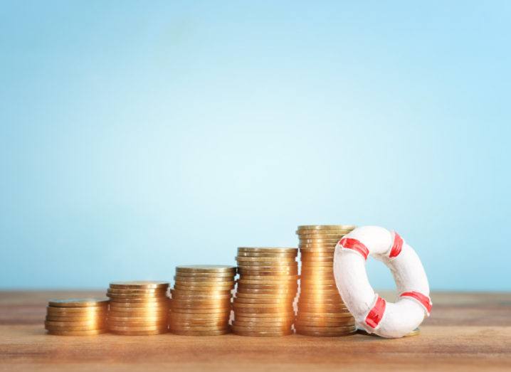 Coins are stacked in a line, with the stacks progressively growing in height. There is a lifebuoy at the end of the coins, balanced against the tallest tower.