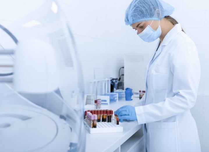 A scientist is standing in a lab and is placing blood samples onto a stand. The stand is a short, white grid and is not yet filled with the vials that are to be tested. The scientist is wearing a mask, gloves, a lab coat and a hair covering, and lab apparatus can be seen in the background.