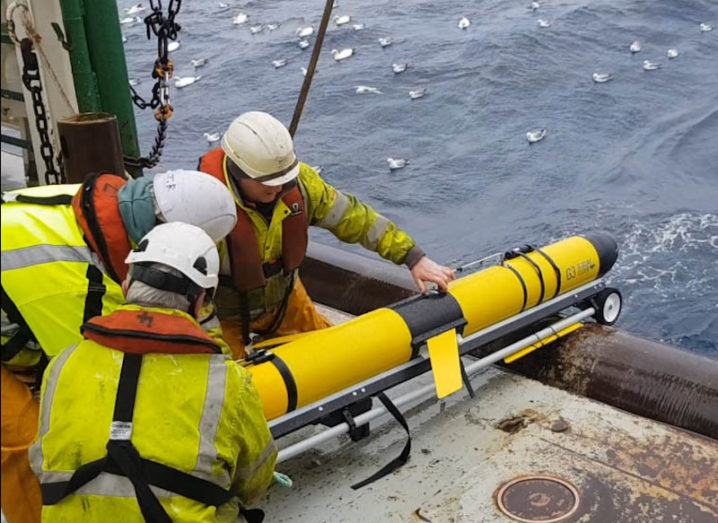 The ocean glider is being deployed by three people from a boat. The glider is a large, yellow tube that is angled towards the water. The people are wearing yellow overalls with life-preserver jackets over them. There are many seagulls in the water.