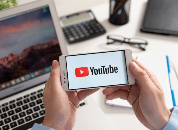 YouTube logo displayed on somebody's phone which they are holding in their hands sitting at a desk in a home office.