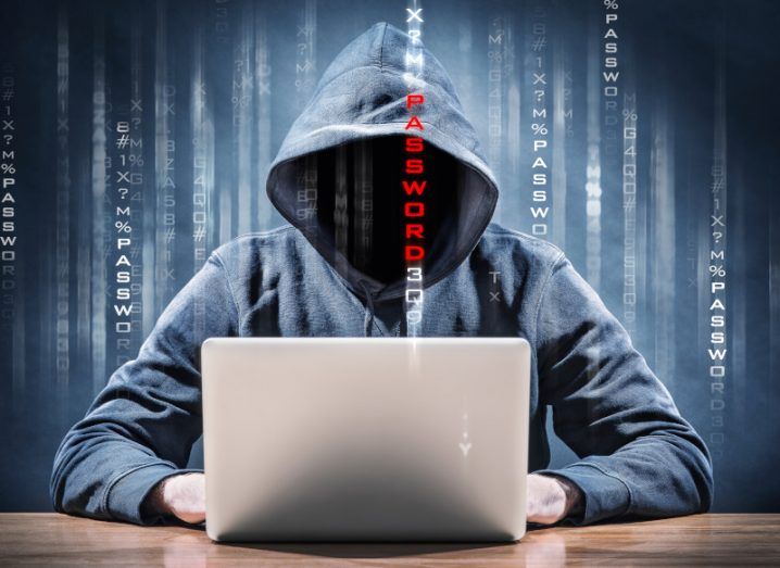 Hooded figure sitting at a table with face obscured by darkness hacking or performing a cyberattack on a computer. Lines of code run vertically down the screen.