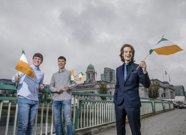 Three young men stand on a bridge in a city, each waving small Irish flags.