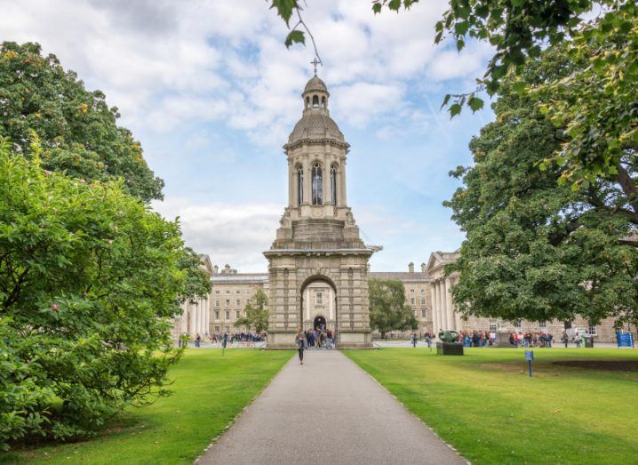 Old bell tower at Trinity College Dublin, with flourishing trees lining the centre path. There are people standing around the tower.