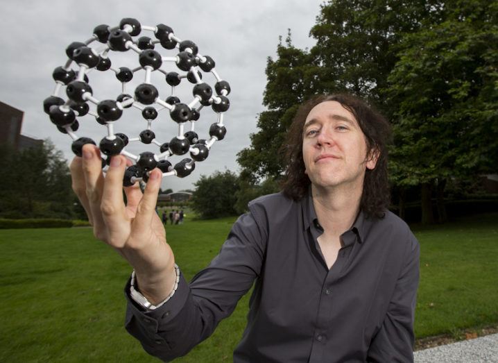 Prof Damien Thompson is pictured holding a model of a molecule. The molecule comprises black plastic atoms connected by small white plastic poles.