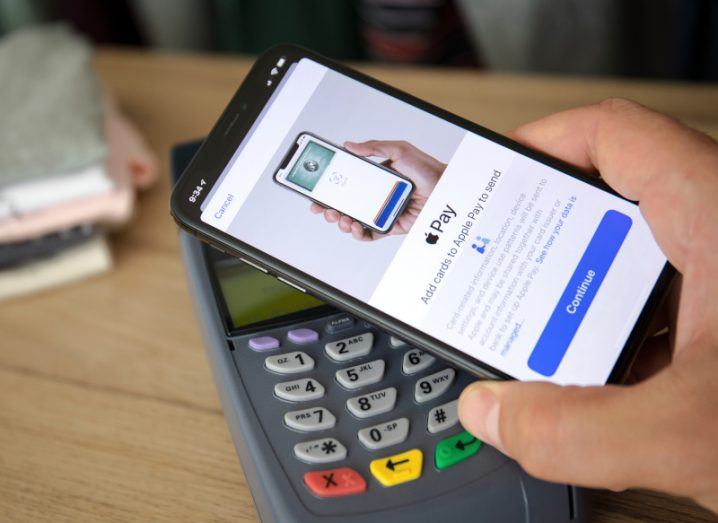 Hand holding a smartphone with Apple Pay's logo above a shop reader.