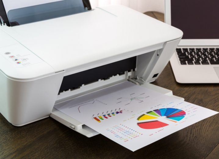 A close-up of a printer with a page of colourful graph sticking out beside a laptop.