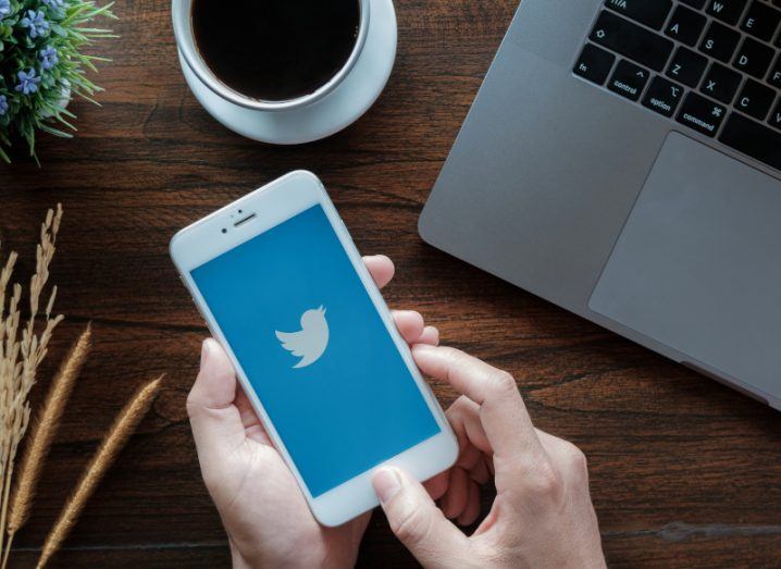 Someone is holding an iPhone with the Twitter logo on the screen over a wooden desk. There is also a computer, cup of coffee and a plant on the desk.