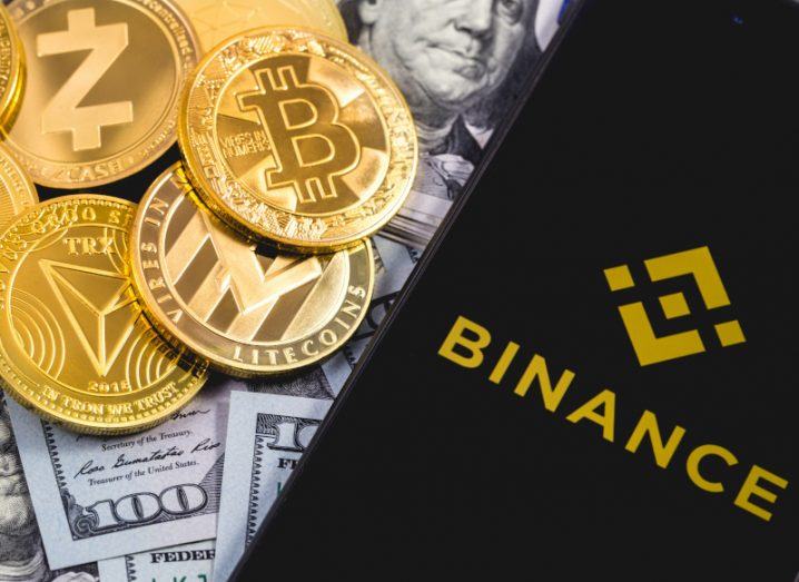 Binance logo is displayed on a smartphone screen, which is resting on a pile of US dollar notes and gold bitcoins.