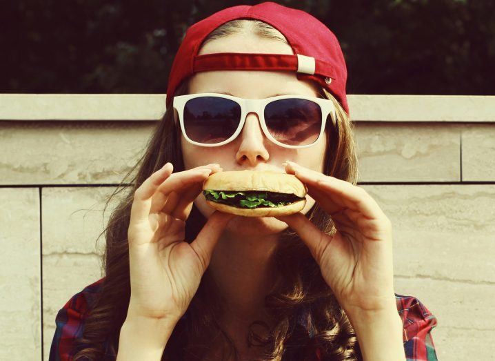 Young woman wearing a backwards baseball cap and sunglasses holding a plant-based burger up to her mouth in a funny pose.