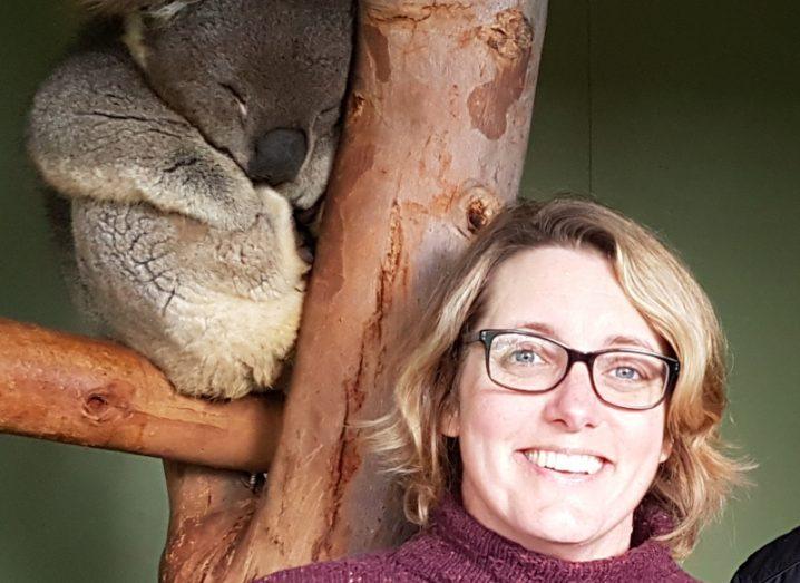 A woman with short blonde hair smiles beside a tree holding a koala bear on the branch.