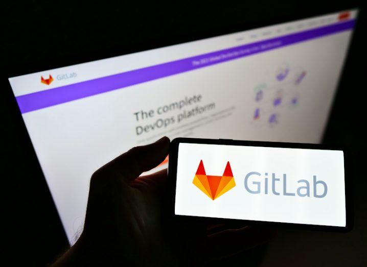 A hand holding a phone with the GitLab logo on it. In the background, GitLab is open on a computer screen, slightly out of focus.