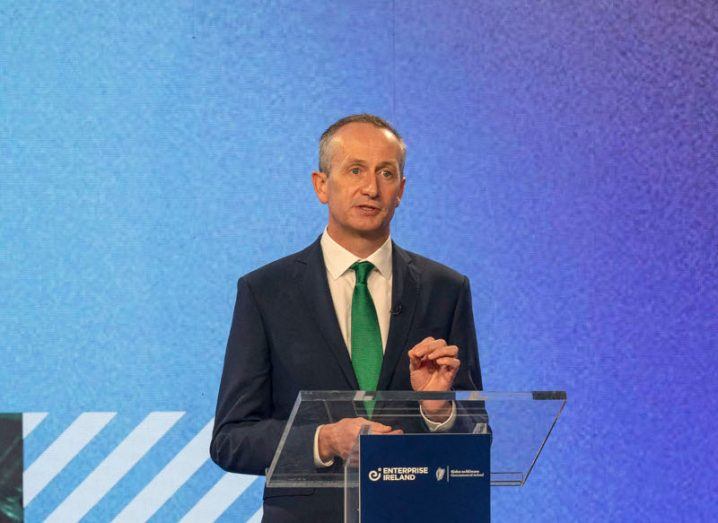 Enterprise Ireland CEO Leo Clancy stands at a clear perspex podium delivering a speech against a textured blue background. He is wearing a dark suit and emerald green tie.