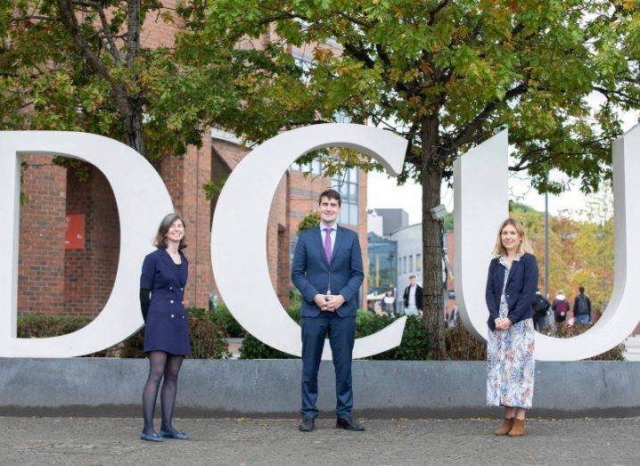 Three people stand in front of a sign that spells out DCU in large, white letters.