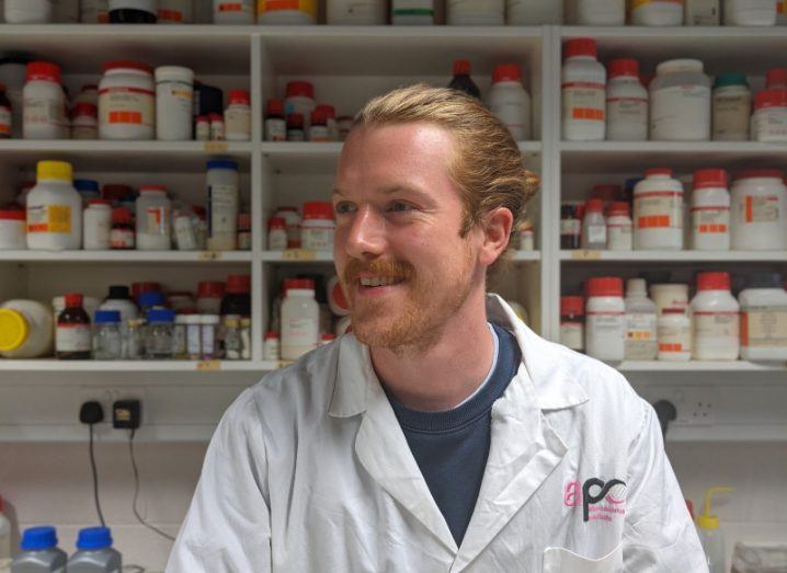 Researcher Jack William Daly in a white lab coat bearing the APC logo. Behind him are shelves filled with labelled containers of various sizes.