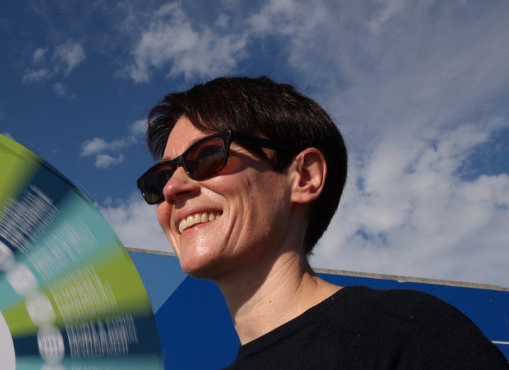 A close-up of a woman with short dark hair smiling off to the left on a sunny day. She is wearing sunglasses against a blue sky.