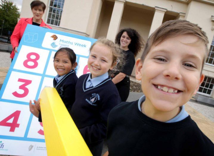 Minister for Education Norma Foley holding a giant board with numbers on it while three schoolchildren smile at the camera and hold a giant yellow pencil.