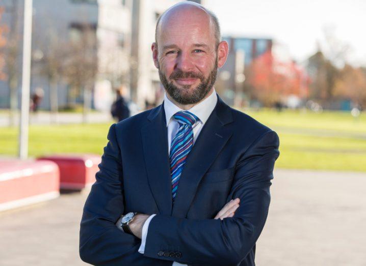 A smiling man in a blue suit and striped tie stands with his arms folded on the campus of Maynooth University.