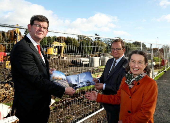 Three people standing outside at a construction site at UCD holding a book of plans and smiling as a digger works in the background.