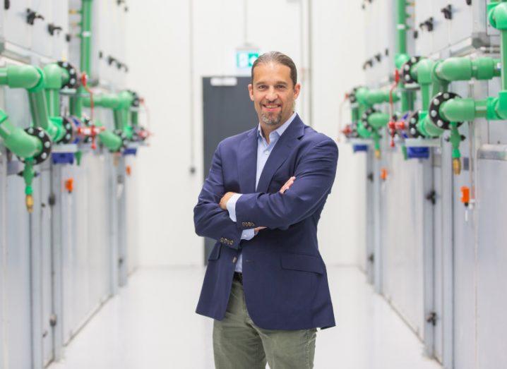 A man in a suit stands in a data centre smiling at the camera.