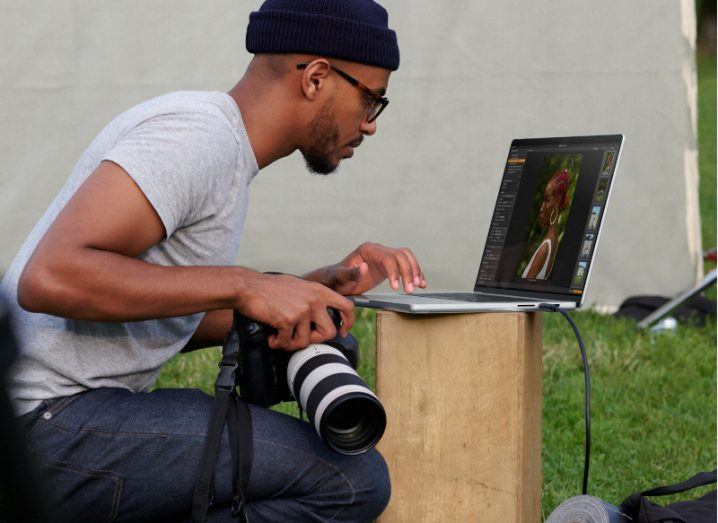 A man sits at an Apple MacBook laptop, while holding a camera in one hand.