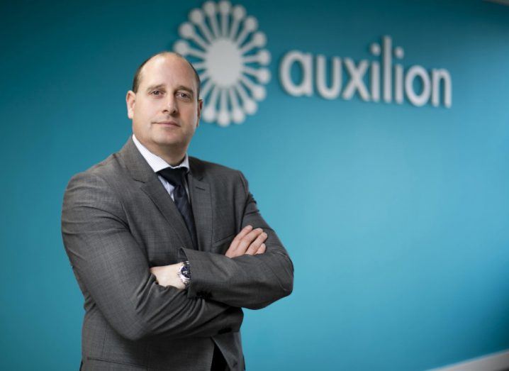 Photo of Auxilion CTO Donal Sullivan standing with arms folded in front of blue wall with the company logo on it.