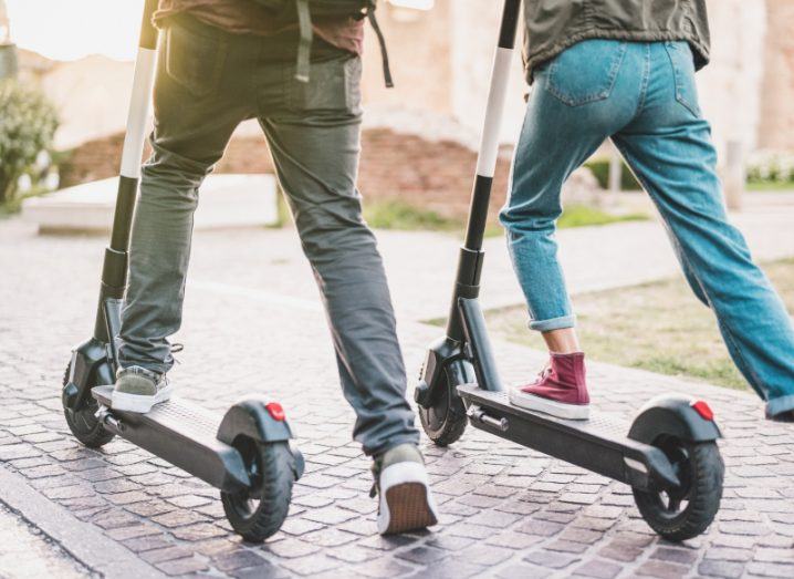 Two people mounted on e-scooters moving forward with the sun in the background.