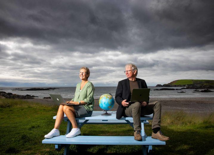 Future Planet co-founders Ingrid De Doncker and Donal Daly sitting at a blue public table with laptops in hand and a globe placed in between them. The sea and mountains can be seen in the background under an overcast dark grey sky.