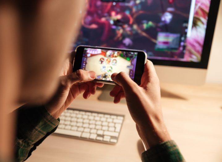 View from above a man's shoulders, who is playing a game on a smartphone in his hands. A desktop screen and keyboard are in the background.