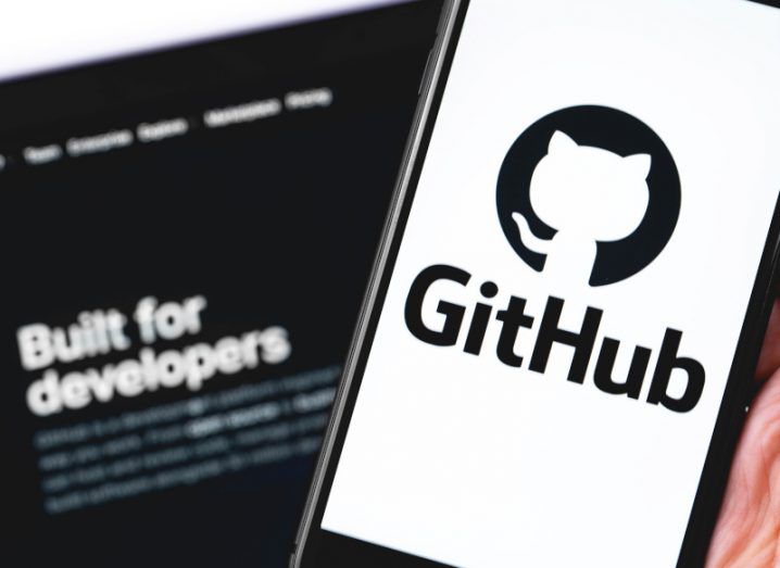 GitHub logo on a smartphone screen with blurred computer screen in the background that reads "built for developers".