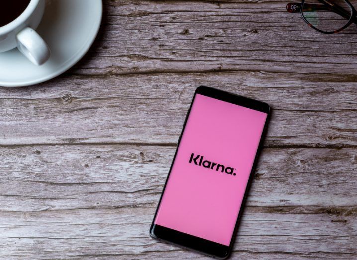 Smartphone with Klarna logo on screen kept on table. A cup of coffee and glasses kept beside the phone.
