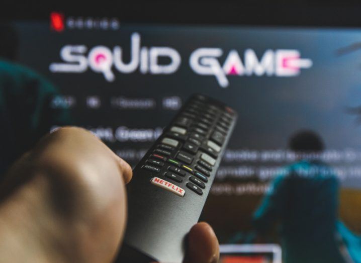 Hand holding a remote control which is pointing at a TV screen showing Squid Game on Netflix. The Netflix logo can also be seen on a remote control.