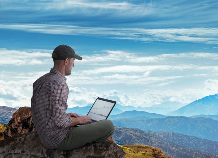 Man working remotely outdoors with laptop. Mountains in the background.