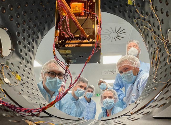 A group of people in protective gear look into a container that is housing the small cube-shaped satellite.