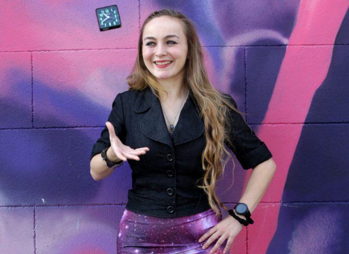 Tammy Strickland, wearing a black blazer and galaxy-print skirt stands in front of a wall graffitied in bright shades of pink and purple.