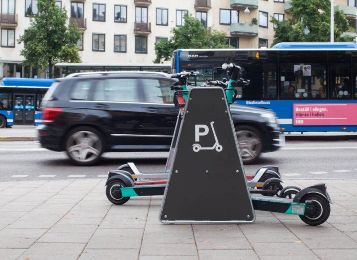 E-scooter parking station with parked vehicles. A moving car is travelling down the street in the background.